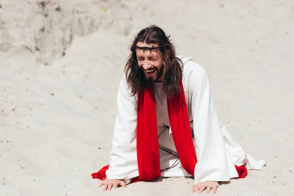 Laughing Jesus in robe, red sash and crown of thorns standing on knees and touching sand with hands in desert — Stock Photo