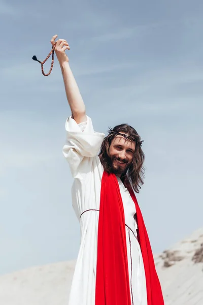 Happy Jesus in robe, red sash and crown of thorns holding rosary in raised hand in desert — Stock Photo