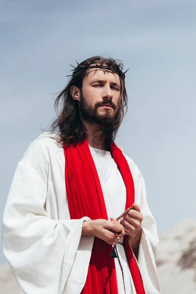 Jesus in robe, red sash and crown of thorns holding rosary and praying with closed eyes in desert — Stock Photo