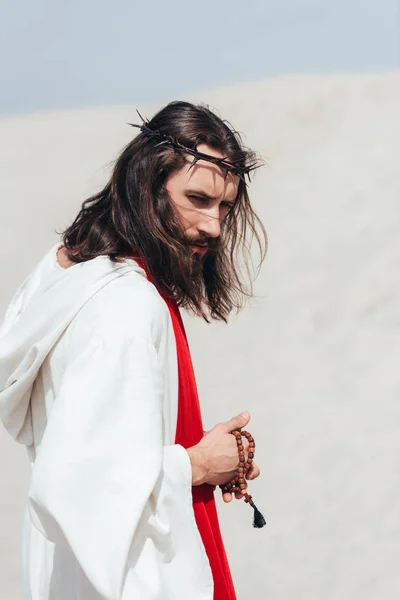 Side view of Jesus in robe and red sash holding wooden rosary in desert — Stock Photo