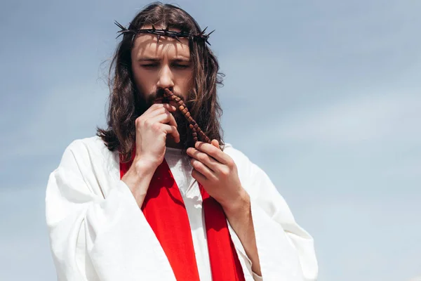 Portrait of Jesus in robe, red sash and crown of thorns kissing rosary against blue sky — Stock Photo