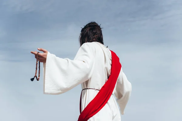 Back view of Jesus in robe, red sash and crown of thorns holding rosary against blue sky — Stock Photo