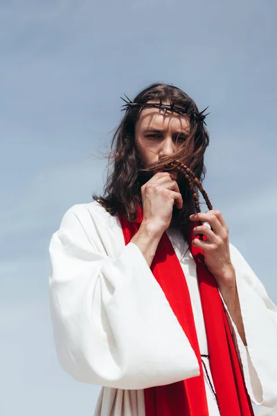 Jésus en robe, ceinture rouge et couronne d'épines embrassant le chapelet contre le ciel bleu — Photo de stock