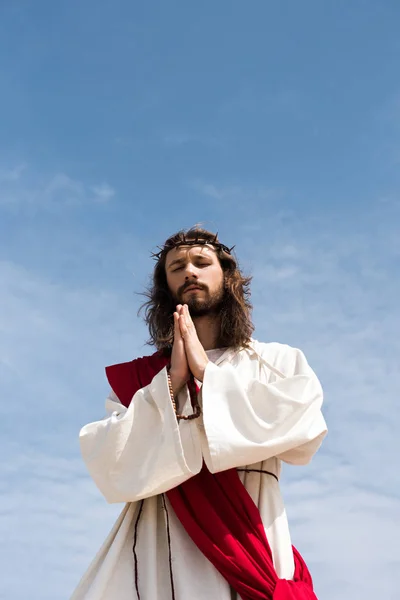 Low angle view of Jesus in robe, red sash and crown of thorns holding rosary and praying with closed eyes against blue sky — Stock Photo