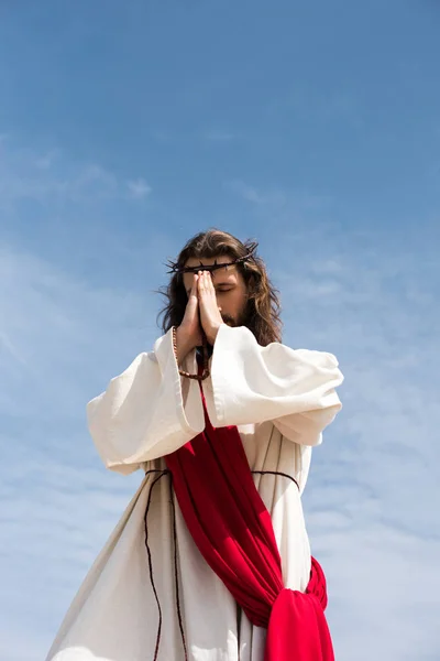 Low angle view of Jesus Christ in robe, red sash and crown of thorns holding rosary and praying against blue sky — Stock Photo