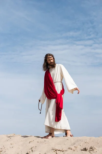 Jesús en túnica, faja roja y corona de espinas sosteniendo el rosario y caminando sobre una colina arenosa en el desierto - foto de stock