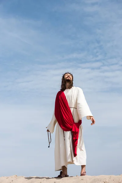 Jesus in robe, red sash and crown of thorns holding rosary and looking up in desert — Stock Photo