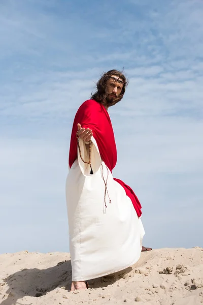 Jesus in robe, red sash and crown of thorns holding rosary and giving hand in desert — Stock Photo