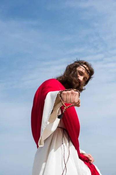 Visão de baixo ângulo de Jesus em roupão, faixa vermelha e coroa de espinhos mostrando punho com rosário contra o céu azul — Fotografia de Stock