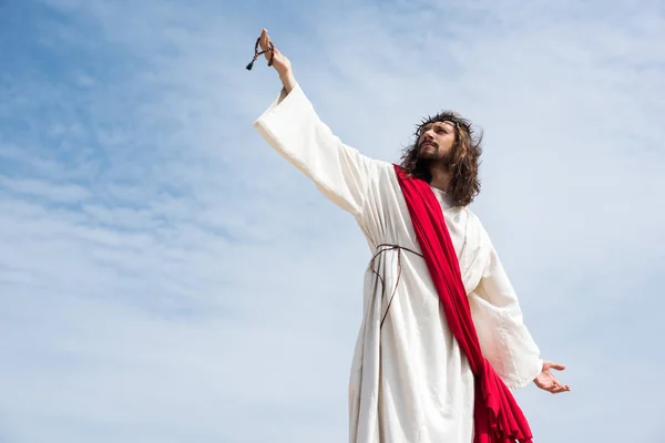 Vista de ángulo bajo de Jesús en túnica, faja roja y corona de espinas sosteniendo rosario en la mano levantada contra el cielo azul - foto de stock