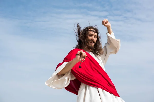 Low angle view of Jesus in robe, red sash and crown of thorns having fun and jumping with rosary against blue sky — Stock Photo