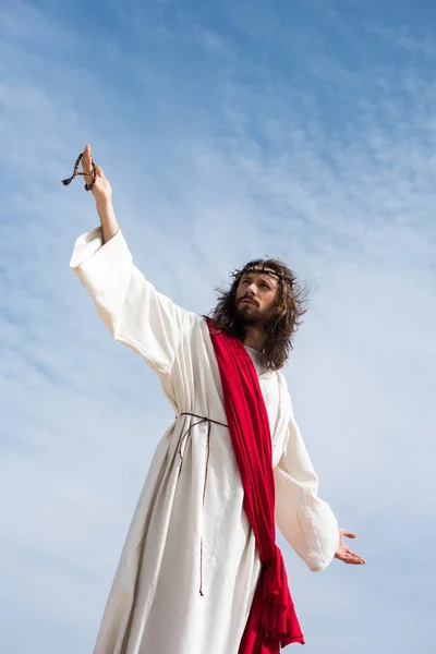 Low angle view of Jesus in robe, red sash and crown of thorns holding rosary and standing with open arms against blue sky — Stock Photo