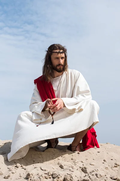 Jesus in robe, red sash and crown of thorns praying with rosary and squatting on sandy hill in desert — Stock Photo