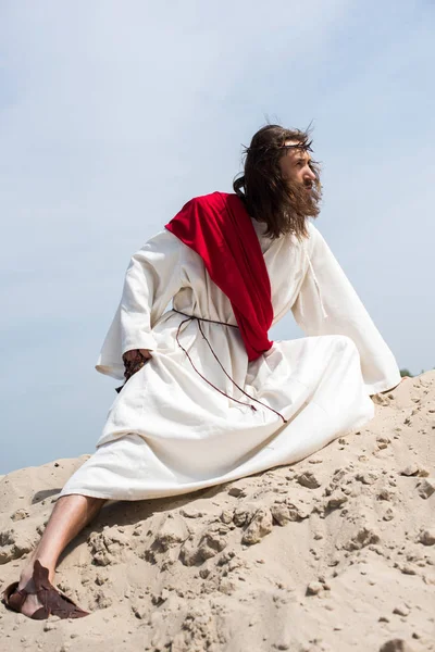 Side view of Jesus in robe, red sash and crown of thorns holding rosary and squatting on sandy hill in desert — Stock Photo