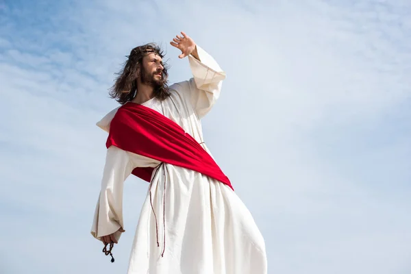Jesus in robe, red sash and crown of thorns holding rosary and protecting face from light in desert — Stock Photo