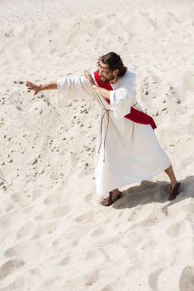 Hochwinkelaufnahme von Jesus in Robe, roter Schärpe und Dornenkrone, der auf Sand steht und in der Wüste gestikuliert — Stockfoto