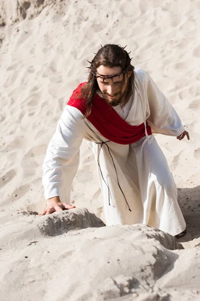 High angle view of Jesus in robe, red sash and crown of thorns climbing sandy hill in desert — Stock Photo
