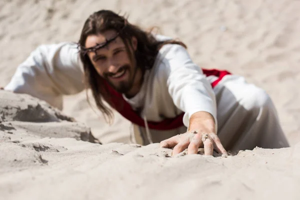 High angle view of smiling Jesus in robe, red sash and crown of thorns climbing sandy hill in desert — Stock Photo