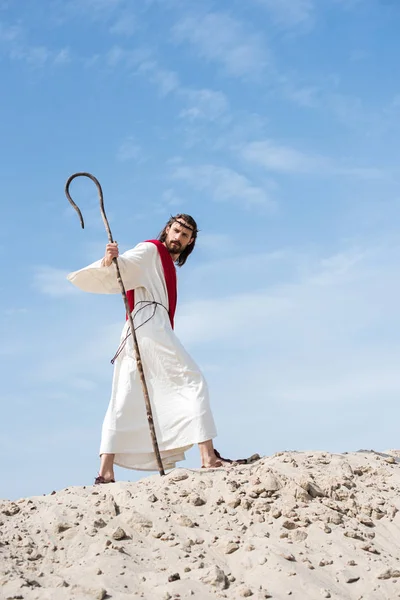 Low angle view of Jesus in robe, red sash and crown of thorns walking on sandy hill with staff in desert — Stock Photo