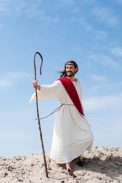 Улыбающийся Иисус в мантии, красной ленте и терновом венце, стоящем с деревянным посохом в пустыне — стоковое фото