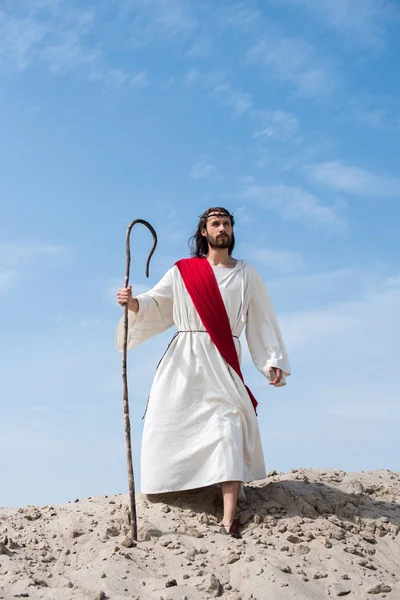 Jesus in robe, red sash and crown of thorns standing on sandy hill with wooden staff in desert — Stock Photo