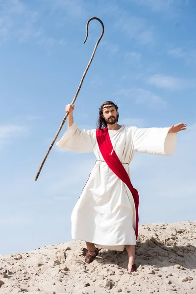 Jesus in robe, red sash and crown of thorns swinging wooden staff in desert — Stock Photo