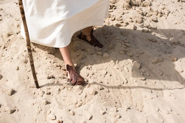 Imagen recortada de Jesús en túnica y sandalias caminando sobre arena con bastón de madera en el desierto - foto de stock