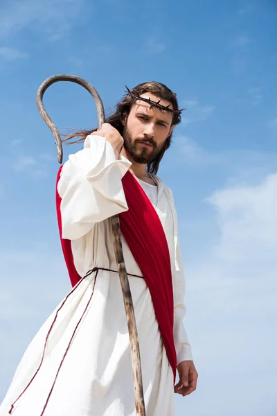 Low angle view of Jesus in robe, red sash and crown of thorns standing with wooden staff in desert and looking at camera — Stock Photo