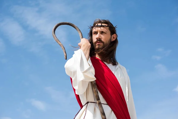 Jesus in robe, red sash and crown of thorns standing with wooden staff against blue sky and looking away — Stock Photo