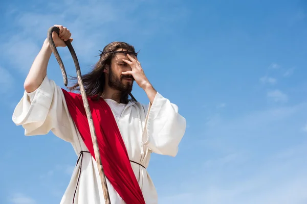 Jesus in robe, red sash and crown of thorns standing with wooden staff in desert and touching forehead — Stock Photo