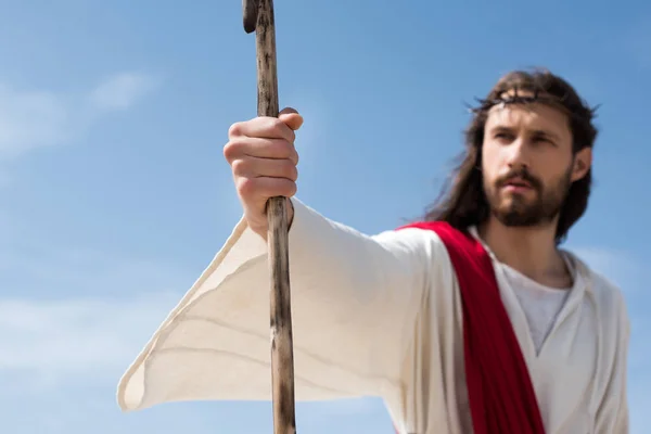 Selective focus of Jesus in robe, red sash and crown of thorns holding wooden staff in desert — Stock Photo