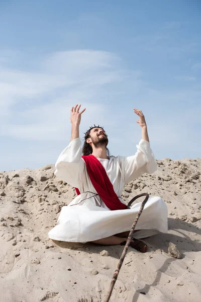 Cheerful Jesus in robe and red sash sitting in lotus position with raised hands and talking with god on sand in desert — Stock Photo