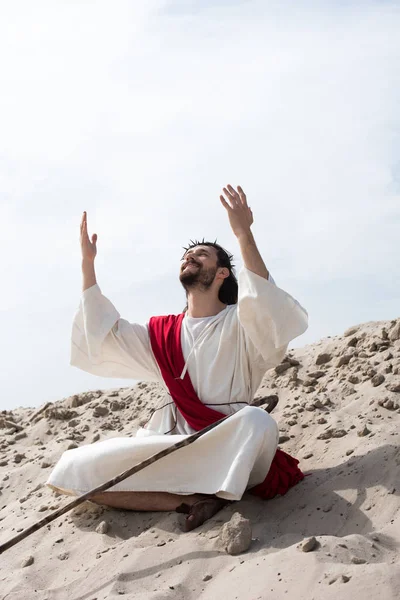 Smiling Jesus in robe, red sash and crown of thorns sitting in lotus position with raised hands and talking with god on sand in desert — Stock Photo