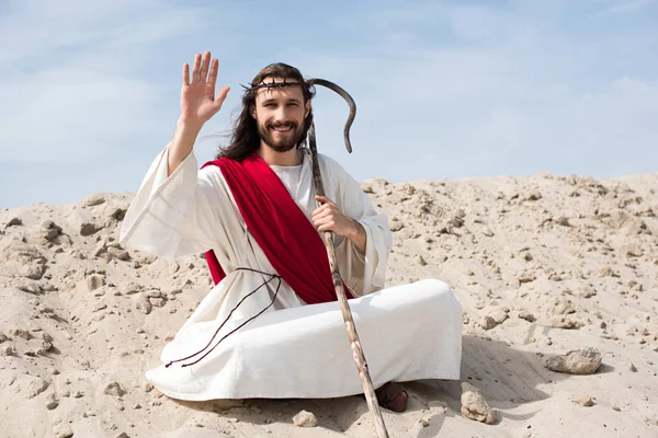 Jesus in robe, red sash and crown of thorns sitting in lotus position on sand in desert and waving hand — Stock Photo