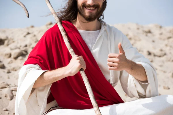 Cropped image of Jesus sitting in lotus position on sand, holding staff and showing thumb up in desert — Stock Photo