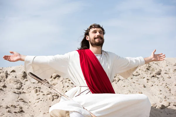 Jesus in robe, red sash and crown of thorns sitting in lotus position with open arms and closed eyes in desert — Stock Photo