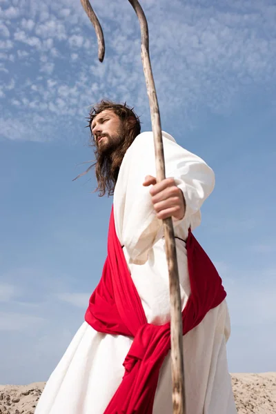 Low angle view of Jesus in robe, red sash and crown of thorns walking in desert with staff — Stock Photo