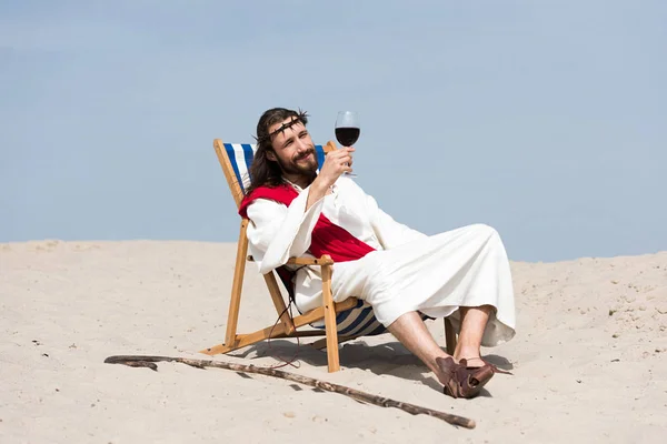 Jésus en robe et ceinture rouge reposant sur une chaise longue et regardant un verre de vin rouge dans le désert — Photo de stock