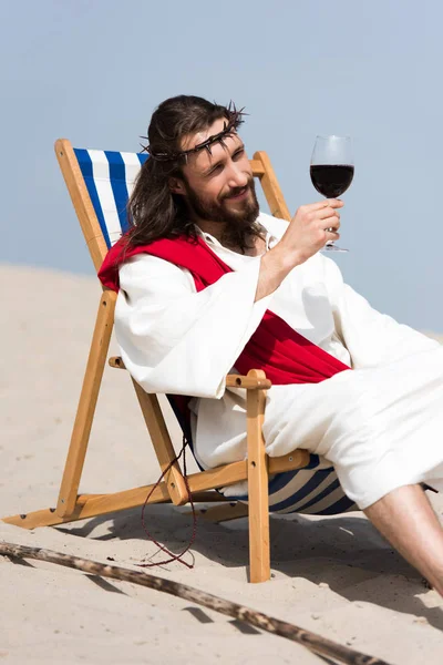 Fröhlicher Jesus in Robe und roter Schärpe, der sich auf einer Sonnenliege ausruht und ein Glas Rotwein in der Wüste betrachtet — Stockfoto