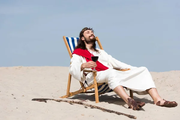 Jesus in Robe und roter Schärpe auf einer Sonnenliege mit einem Glas Rotwein in der Wüste — Stockfoto