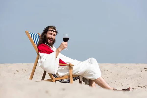 Улыбающийся Иисус в халате и красной ленте, лежащий на шезлонге с бокалом красного вина в пустыне и смотрящий в камеру — стоковое фото