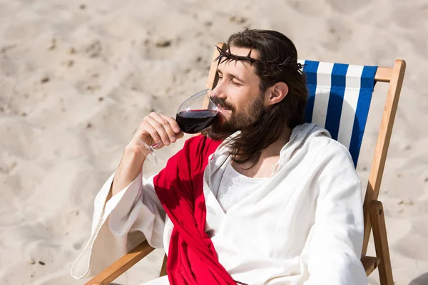 Jesus de roupão e faixa vermelha descansando na espreguiçadeira e bebendo vinho tinto no deserto — Fotografia de Stock