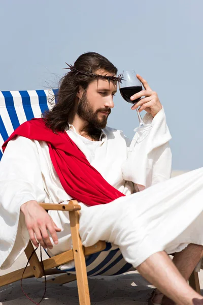 Pensive Jesus in robe and red sash sitting on sun lounger with glass of red wine in desert — Stock Photo