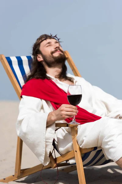 Selektiver Fokus des fröhlichen Jesus in Robe und roter Schärpe auf einer Sonnenliege mit einem Glas Rotwein in der Wüste — Stockfoto