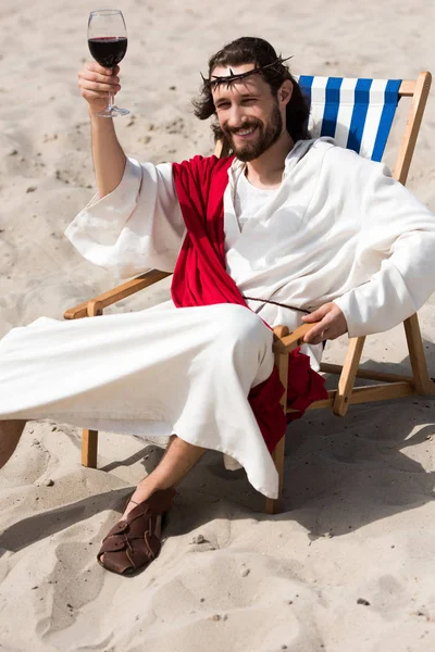Jésus joyeux en robe et ceinture rouge reposant sur une chaise longue et montrant un verre de vin rouge dans le désert — Photo de stock