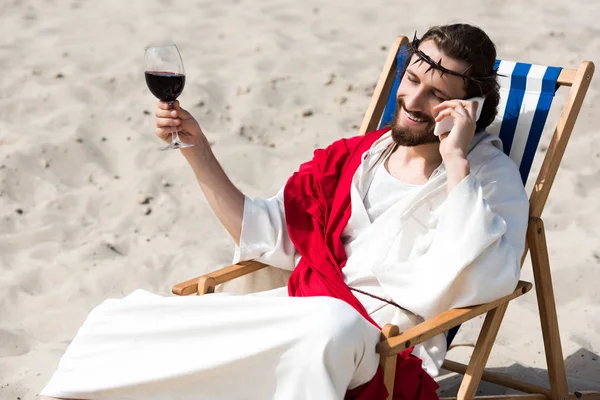 Улыбающийся Иисус отдыхает на шезлонге с бокалом вина и разговаривает со смартфоном в пустыне — стоковое фото