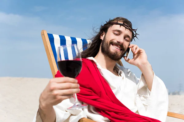 Glücklicher Jesus, der sich mit einem Glas Wein auf einer Sonnenliege ausruht und in der Wüste mit dem Smartphone spricht — Stockfoto