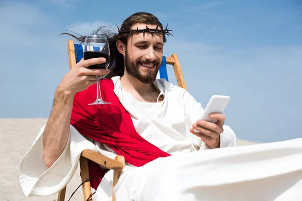 Улыбающийся Иисус отдыхает на шезлонге с бокалом вина и использует смартфон в пустыне — стоковое фото