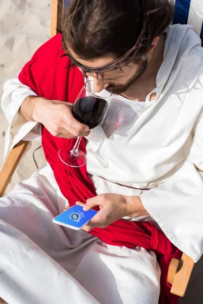 High angle view of Jesus resting on sun lounger, drinking wine and using smartphone with Shazam appliance in desert — Stock Photo