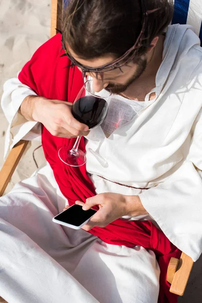 Vista de ángulo alto de Jesús descansando en una tumbona, bebiendo vino y usando un teléfono inteligente con pantalla en blanco en el desierto - foto de stock